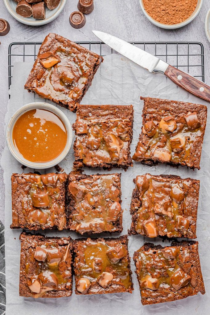 Fudgy Rolo Brownies (Easy, No-Mixer, Scratch Brownies!) Nine chocolate brownies topped with caramel sauce are arranged on parchment paper over a cooling rack, with a bowl of caramel and a knife nearby.