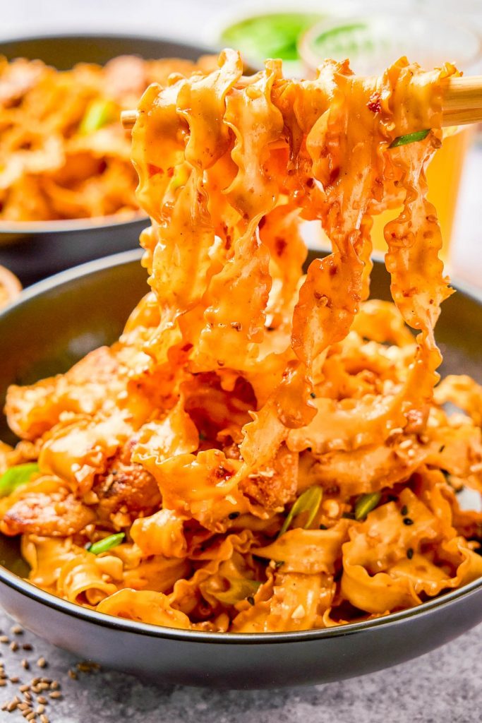 The Best Better-Than-Takeout Recipes (20 Copycat Asian Recipes!) A bowl of spicy, saucy noodles with pieces of meat and green onions, being lifted by chopsticks.